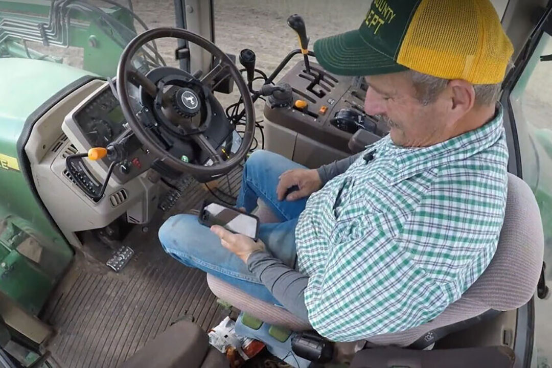 Boer checkt zijn smartphone terwijl zijn tractor gedeeltelijk autonoom rijdt dankzij AgJunction autosteer. - Foto: Still uit AgJunction-video